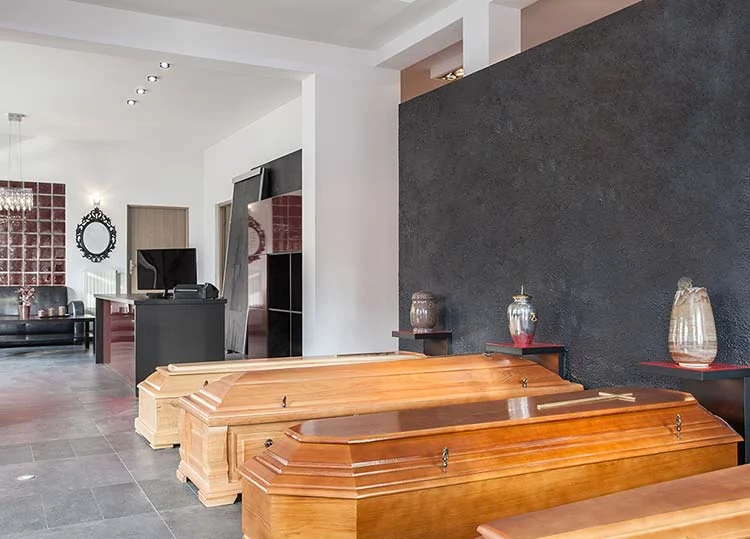 Ein modernes Bestattungsinstitut mit Holzsärgen auf dem Boden, Urnen in Regalen, einer schwarz strukturierten Wand, einer Sitzecke und einem Schreibtisch mit einem Computer im Hintergrund - betrieben von Geprüfte Bestatter für vertrauenswürdige Dienstleistungen. - Bestattungsunternehmen in Winsen, Harburg und in der Elbmarsch.