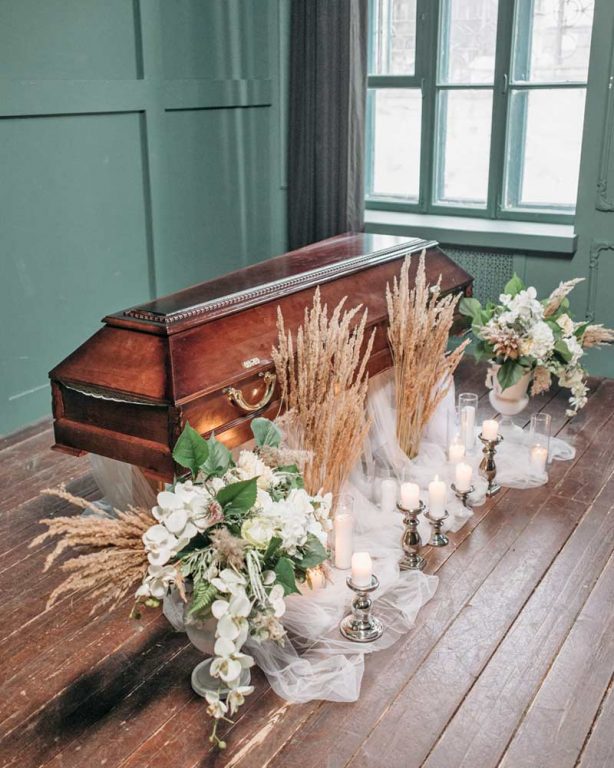 Ein Holzsarg ist von weißen Blumen, getrockneten Gräsern, brennenden Kerzen und durchsichtigen Stoffen umgeben und stellt einen der vielen Bestattungsarten in einem Raum mit grün getäfelten Wänden und großen Fenstern dar. - Bestattungsunternehmen in Winsen, Harburg und in der Elbmarsch.