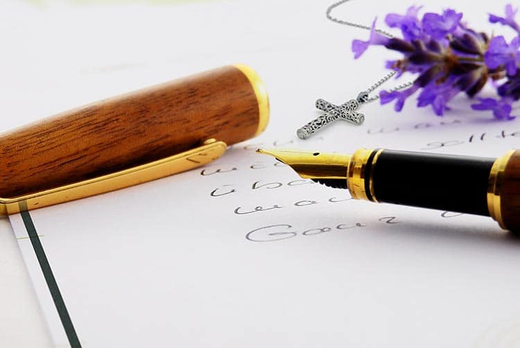 Nahaufnahme eines Füllfederhalters mit Holzkappe auf handgeschriebenem Papier, daneben Beileidstexte, eine silberne Kreuzkette und violette Blumen. - Bestattungsunternehmen in Winsen, Harburg und in der Elbmarsch.