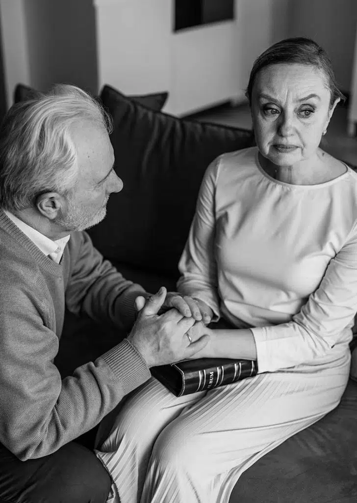 Ein älterer Mann hält sanft die Hände einer älteren Frau, die neben ihm auf einer Couch sitzt. Die Frau sieht ergriffen aus und hält ein geschlossenes Buch in ihrem Schoß. Die Szene wirkt ernst und unterstützend - ein Schwarz-Weiß-Foto, das Momente nach einem Todesfall widerspiegelt. - Bestattungsunternehmen in Winsen, Harburg und in der Elbmarsch.