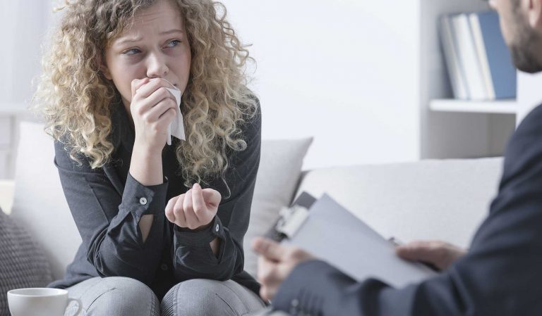 Eine Frau mit lockigem Haar sitzt auf einer Couch, sieht aufgeregt aus und hält sich ein Taschentuch vor den Mund, während sie eine einfühlsame Trauerbegleitung mit einer Person führt, die ein Klemmbrett hält, was auf ein Trauergespräch hindeutet. - Bestattungsunternehmen in Winsen, Harburg und in der Elbmarsch.