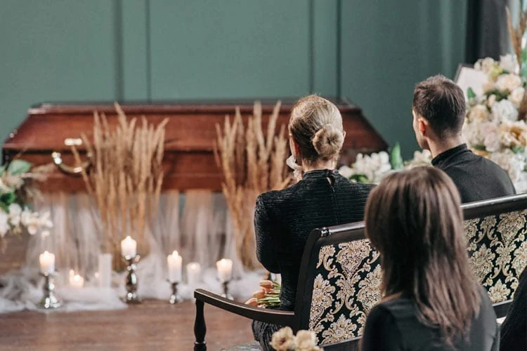 Drei Personen sitzen auf einer Bank vor einem geschlossenen Sarg, umgeben von Kerzen, Blumen und Ziergräsern, und nehmen still an einer Trauerfeier in einem gedämpft beleuchteten Raum teil. - Bestattungsunternehmen in Winsen, Harburg und in der Elbmarsch.
