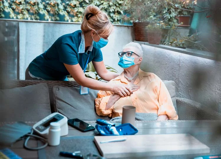 Eine maskierte Pflegerin hilft einer älteren Frau, der ebenfalls maskiert auf einer Couch im Freien in Winsen sitzt. Medizinisches Material und ein Blutdruckmessgerät liegen auf dem Tisch, was auf eine mitfühlende Sterbebegleitung hinweist. - Bestattungsunternehmen in Winsen, Harburg und in der Elbmarsch.