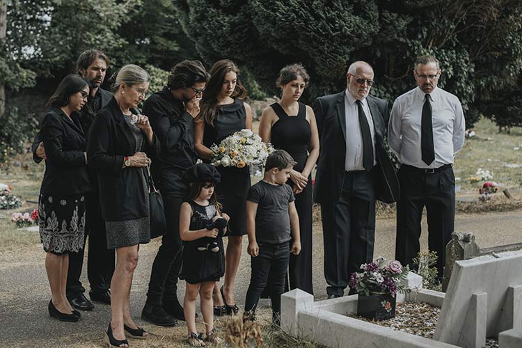 Eine Gruppe schwarz gekleideter Menschen, darunter Erwachsene und Kinder, stehen feierlich um ein Grab mit Blumen auf einem Friedhof in Winsen und trauern um einen Verlust während der Beerdigung. - Bestattungsunternehmen in Winsen, Harburg und in der Elbmarsch.