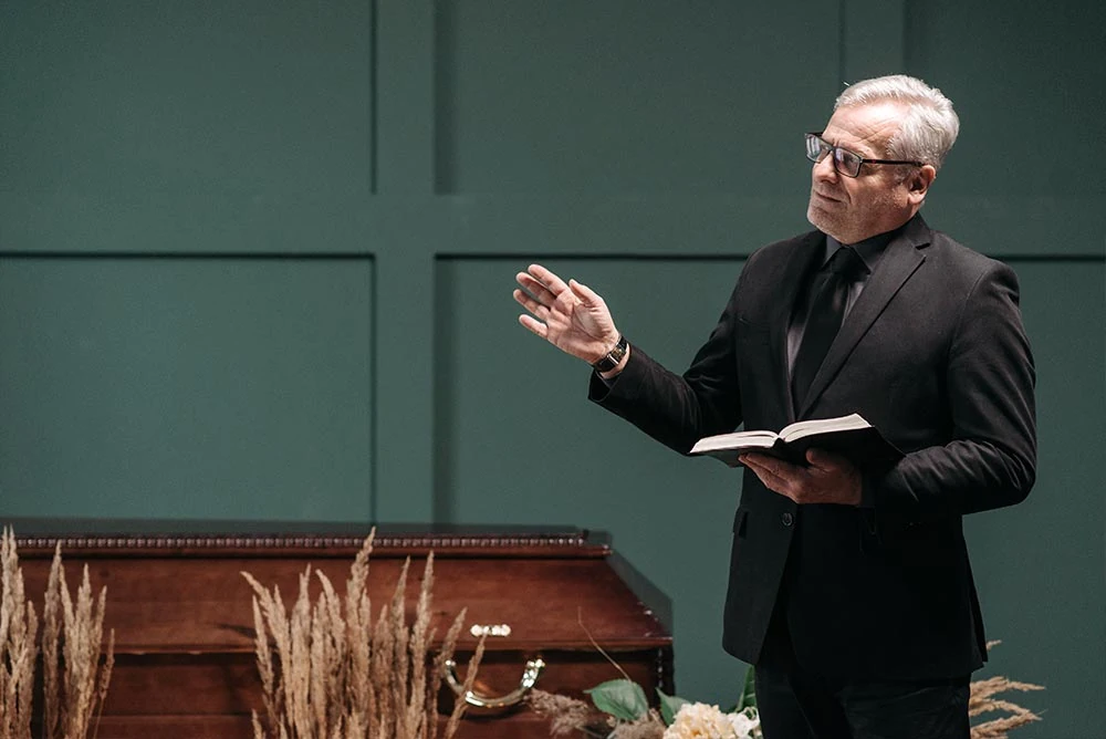 Ein Trauerredner im schwarzen Anzug und mit Brille steht neben einem Holzsarg, hält ein aufgeschlagenes Buch in der Hand und hält eine herzliche Trauerrede bei der Beerdigung. Neben dem Sarg liegen getrocknete Blumengestecke. - Bestattungsunternehmen in Winsen, Harburg und in der Elbmarsch.